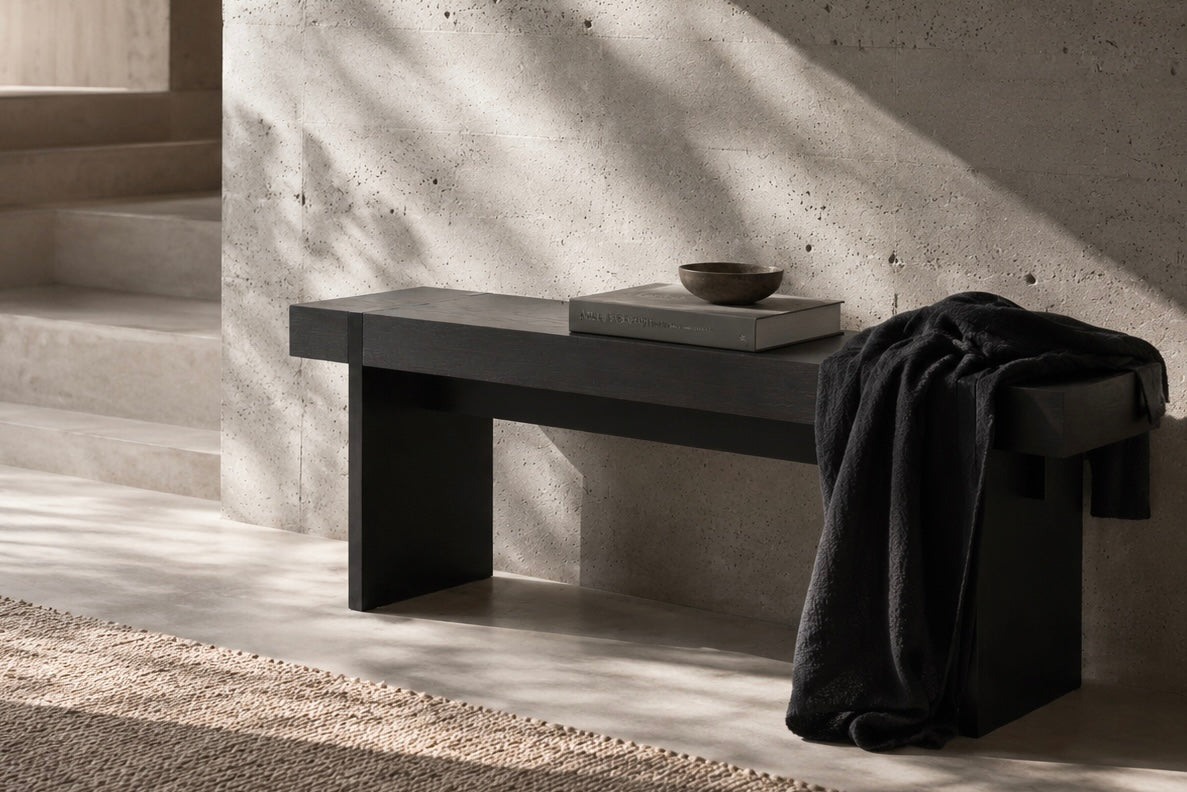 Modern blackened oak bench against a concrete wall with a bowl and book on it.